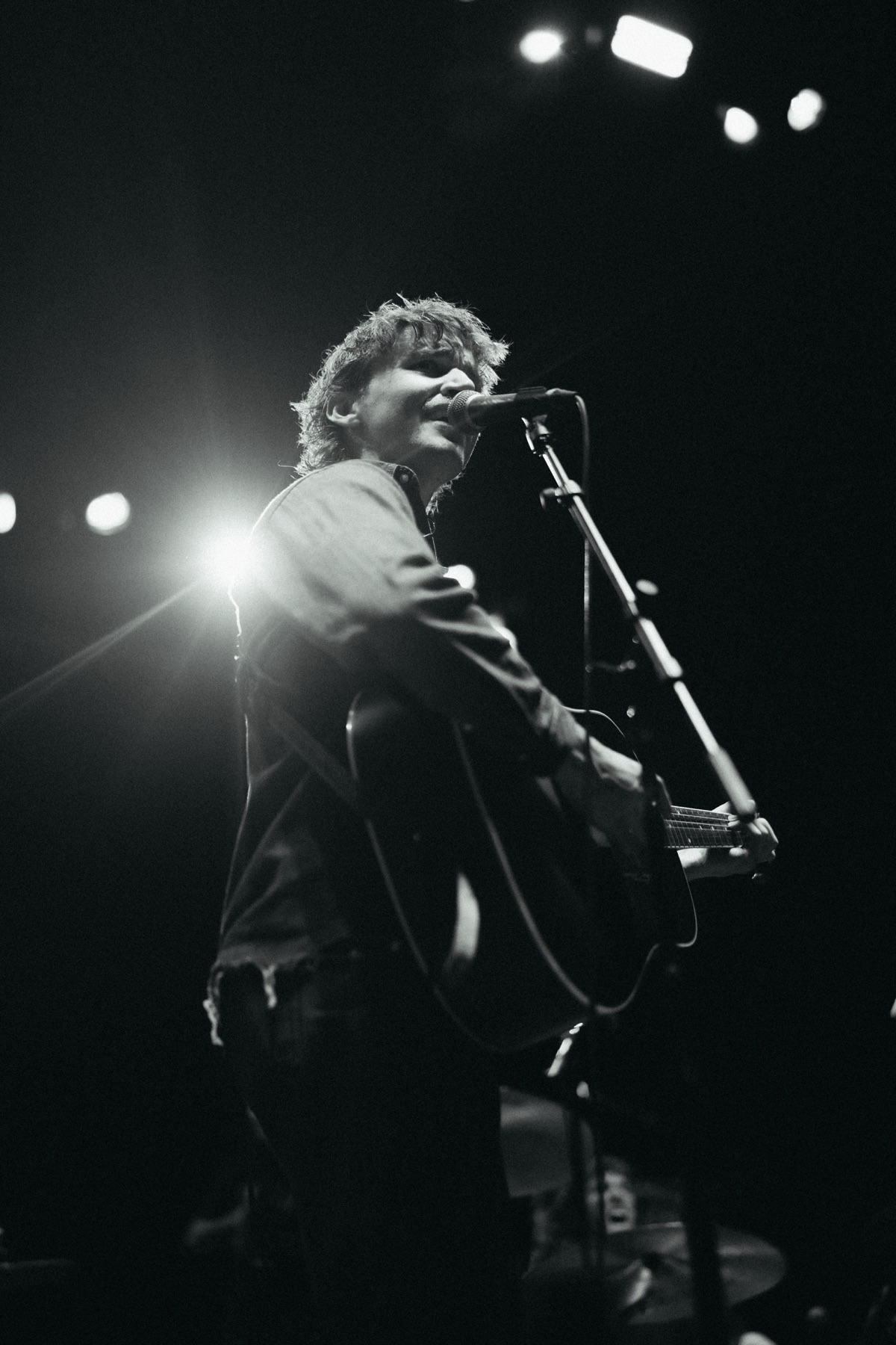 Phil Kane singing into a microphone with his guitar under bright stage lights